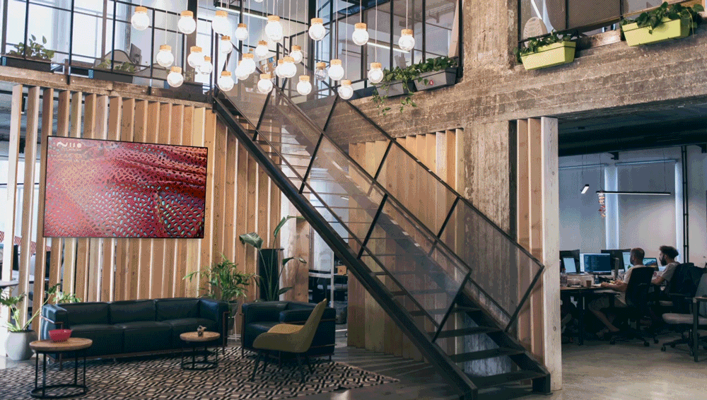 Lobby and entrance of a beautiful public space, with innovative interior design, stairways and couches, and a large screen with the digital artwork: Geist_xyz by ZEITGUISED Powered by Niio